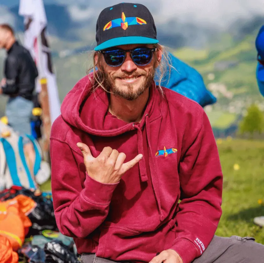 Karlis Jaunpetrovics in a Flying Karlis cap and hoodie making a shaka sign at a paragliding launch site, with mountains behind